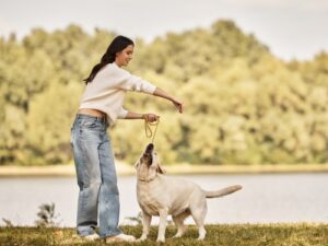 Eine junge Frau in gemütlicher Herbstkleidung spielt fröhlich mit ihrem Hund in der Nähe eines ruhigen Sees.